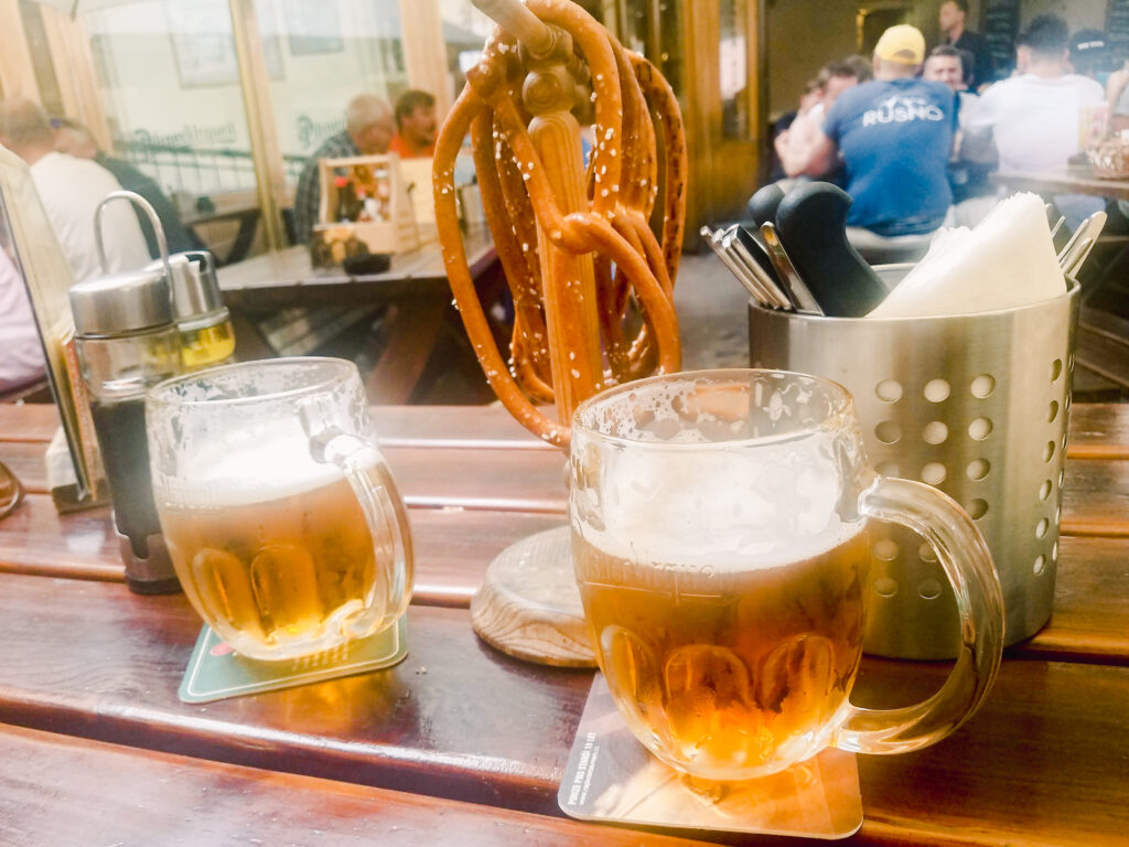 Two large mugs of beer and a pretzel stand on a table in Prague. Drinking beer is definitely one of the best things to do in Prague.