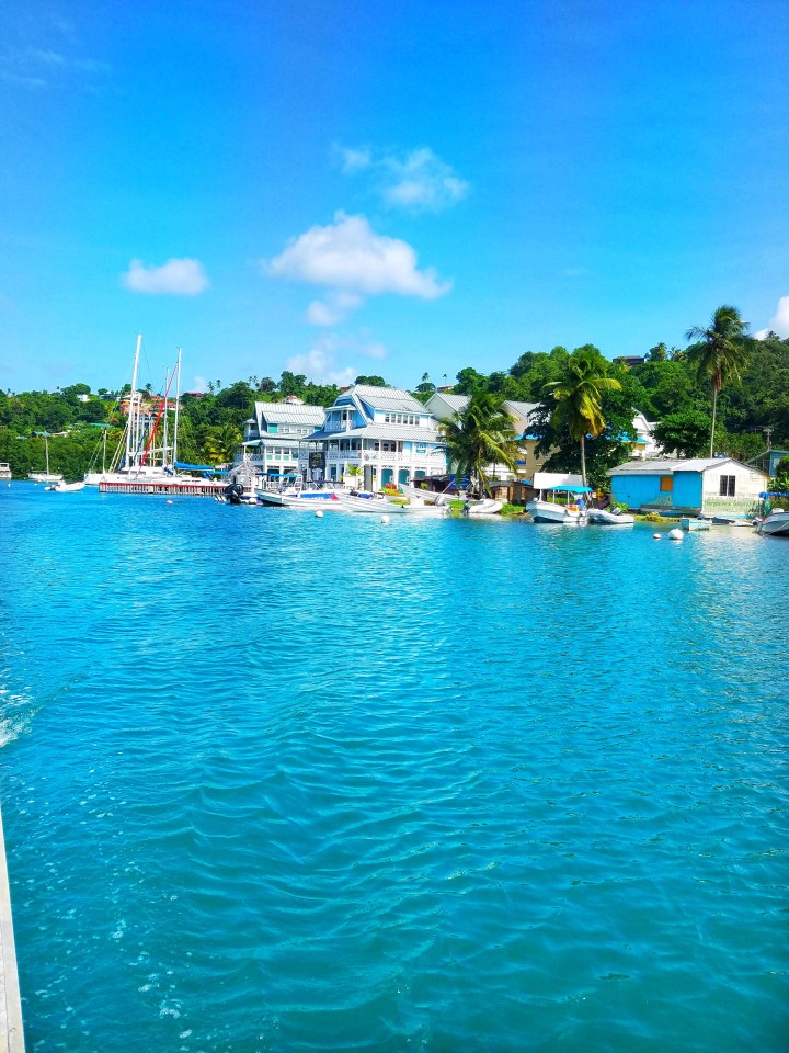 A view of clear blue water, colorful blue houses, and lush green palm trees and mountains. A beautiful view as I sail through St Lucia with a friend. 