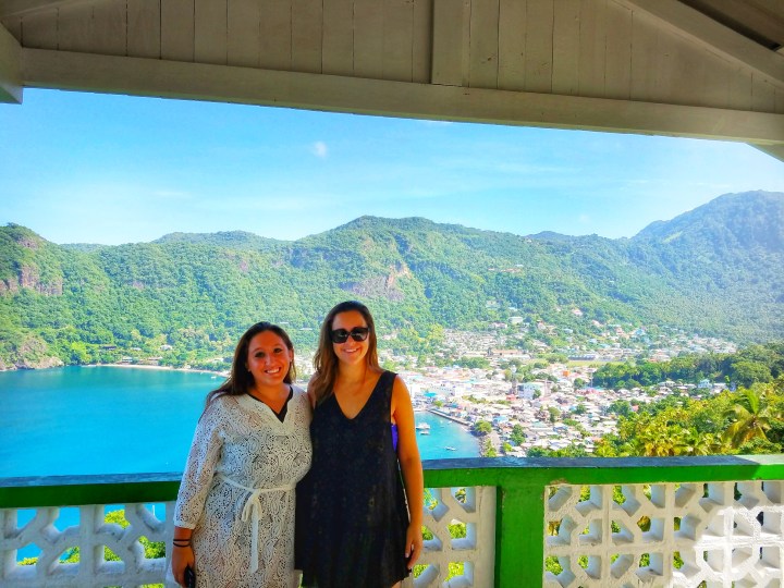 Two girls stand smiling at the camera with mountains, a bay, and colorful houses in the background during a one day trip to Soufriere 