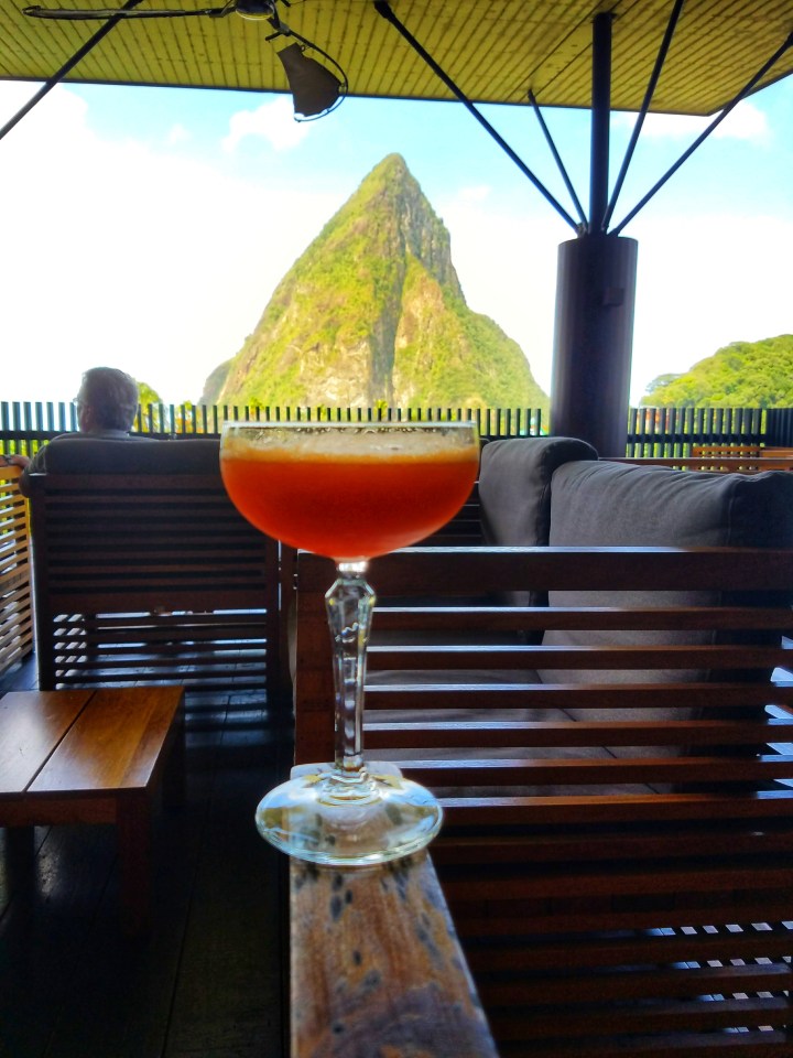 A view of St Lucia's pitons from Boucan restaurant by Hotel Chocolat. There is a fruity cocktail positioned in front of the Pitons view. You must see this view even if you only have one day in Soufriere. 
