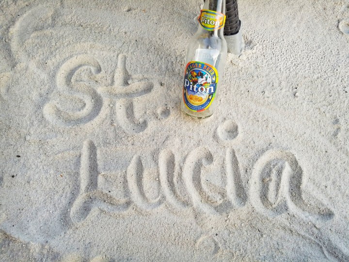 St Lucia written in the sand, and a bottle of St Lucia's local beer on the ground next to it