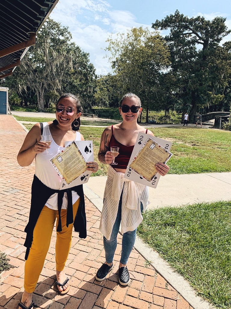 A woman in a white tank top and yellow pants and a girl in a red tank top and jeans hold glasses of champagne, giant playing cards, and descriptions of their birthday predictions from the Champagne Destiny Reading. 