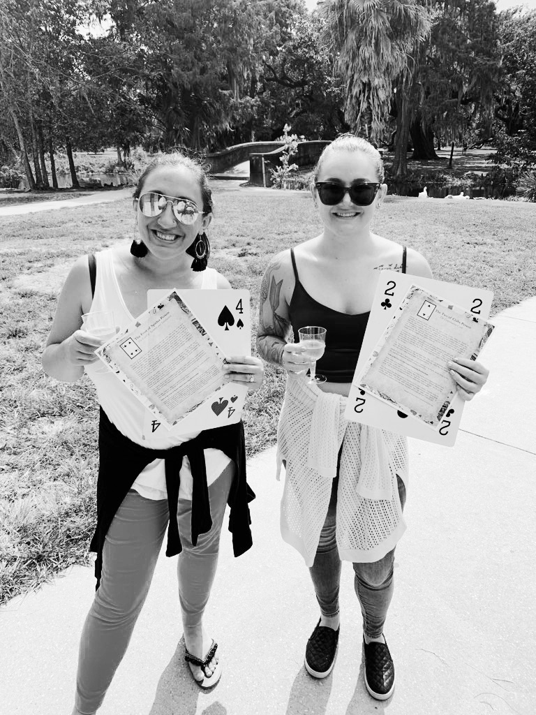 Two women holding giant playing cards. Four of spades is on the left, two of clubs is on the right. Women hold descriptions of their birth date predictions from the Champagne Destiny Reading. 