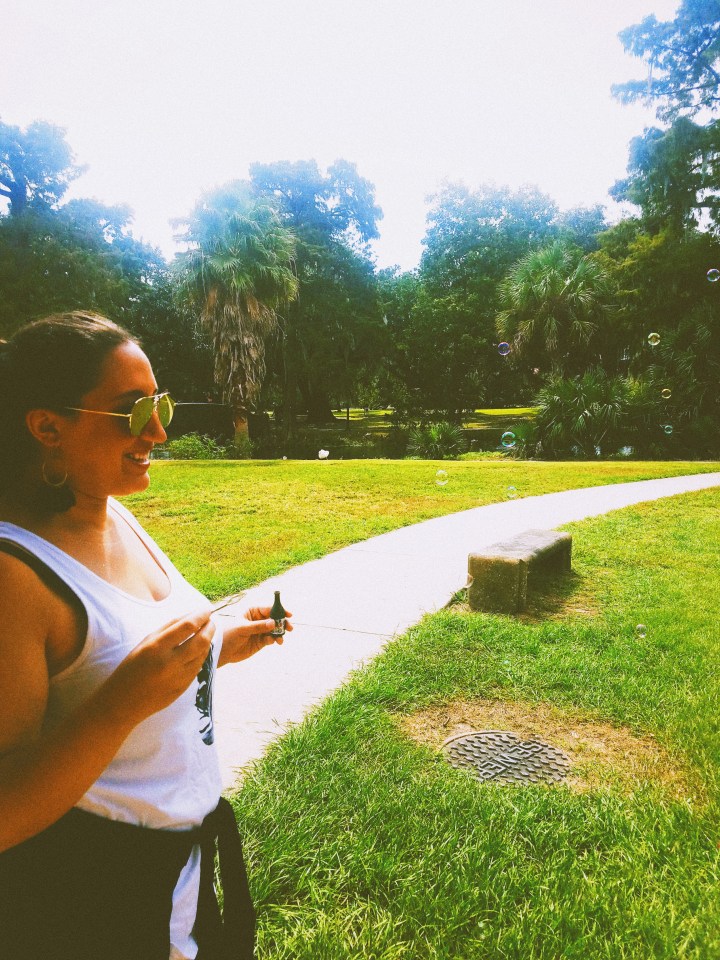 A woman in black feather earrings, a white tank top, and sunglasses blowing bubbles in a park as part of the Champagne Destiny Reading.