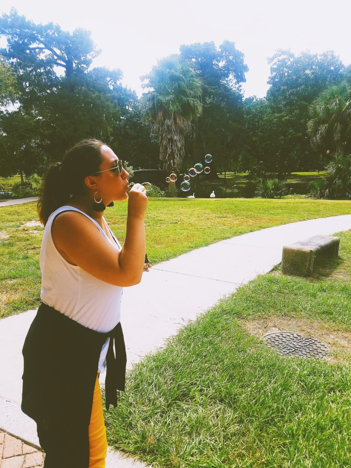 A woman in sunglasses and a white tanktop and feather earrings blows bubbles at the park at the end of the Champagne Destiny Reading. She is wearing yellow pants and a blank sweater tied around waist. 