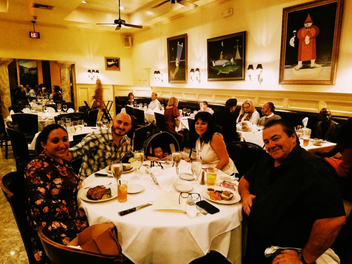 A family sits for dinner in New Orleans. From left to right: a young woman in a floral purple dress, a young man in a plaid shirt, a baby in a stroller, a woman in a white shirt, a man in a black shirt.