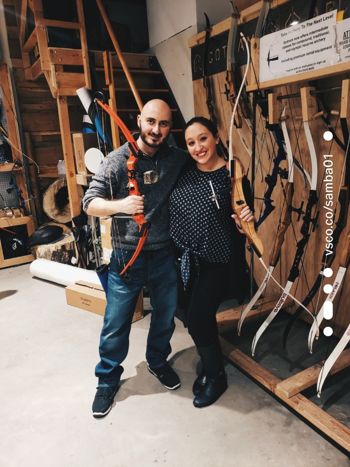 A man in a gray sweater, sneakers, and jeans holds an archery bow while a woman in a polka dot shirt, black jeans, and boots holds a more traditional type of archery bow at Gotham Archery in Brooklyn