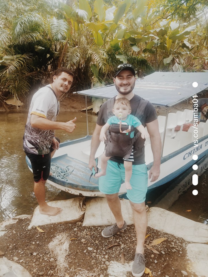 A father has his baby strapped to his chest and is about to climb on board for a monkey boat tour in Costa Rica. A tour guide stands next to him giving a thumbs up before they board. 