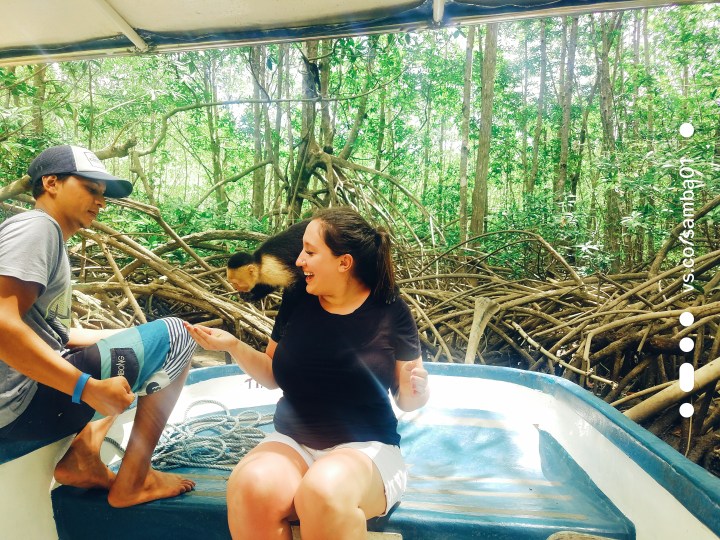 A woman is taking a monkey boat tour in Costa Rica. She sits on the edge of a boat while a monkey climbs onto her back. She has a black t-shirt on and white shorts. The monkey is small, furry, and black and white.
