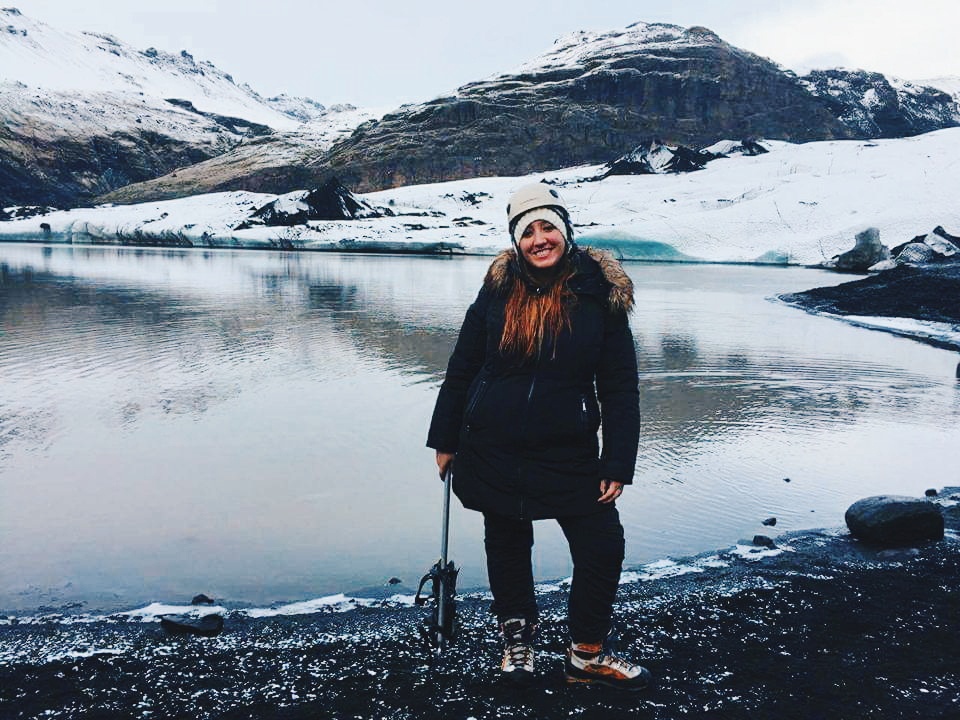 A woman poses in climbing gear next to a lake at Solheimajokull Glacier