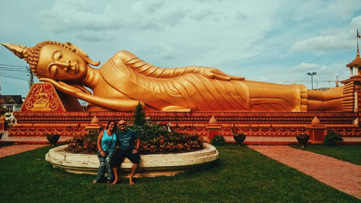 A giant golden reclining, or "sleeping" Buddha outside of a temple in Vientiane, Laos. A couple poses in front of it, sitting on a bench. 