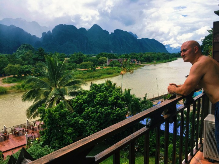 A shirtless bald man looking out over a river with mountains and palm trees in the background in Laos