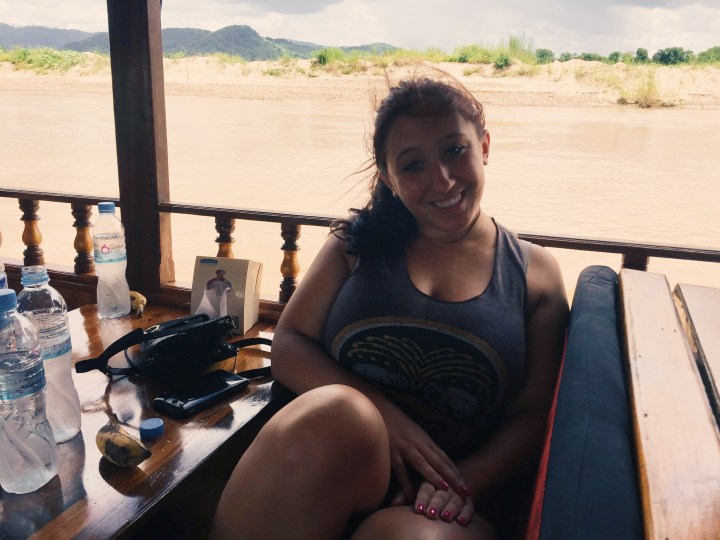 A woman smiles at the camera wearing a gray tanktop and pony tail. She had tan skin and brown hair. She is on board a slow boat that is sailing down the Mekong river. 