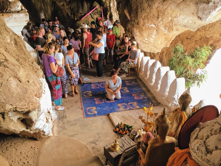 A woman kneels on a prayer mat in the Pak Ou caves. She is shaking a cup full of numbered sticks with an altar in front of her. A group of people wait behind her and look on. 