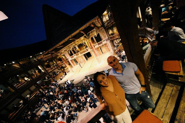 A couple poses for a picture in the gallery of the Globe Theater. The woman has long brown hair and is in a salmon colored shirt and white pants. The man is bald and has a blue t-shirt and jeans. 