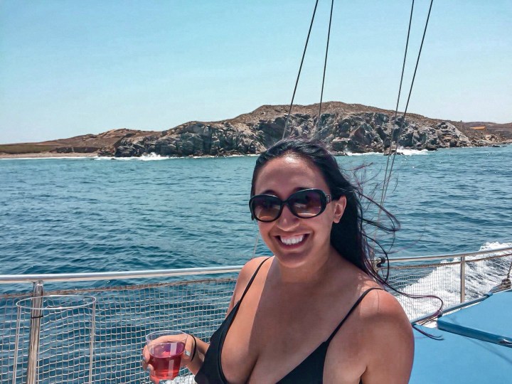 A woman holds a plastic glass full of rose, is drunk and sailing around Mykonos and smiles at the camera wearing sunglasses. 