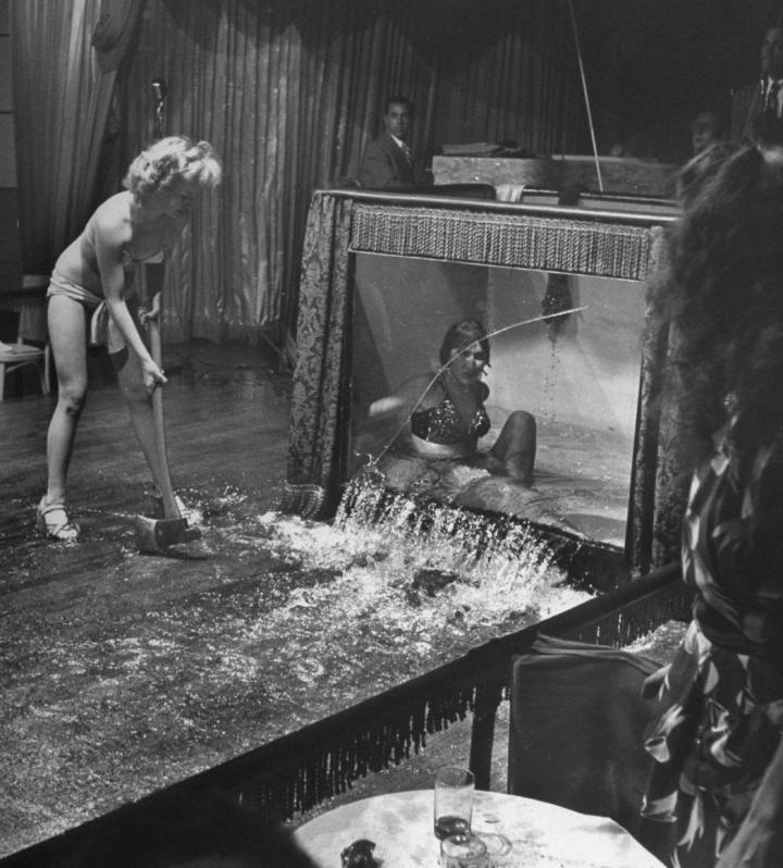 Evangelina smashes a water tank with performer inside. She stands on stage in bikini and heels holding a hammer. Water pours from tank and girl inside of tank appears to be in shock.