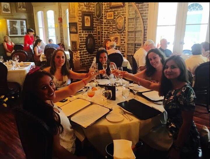 Five girls sit around a circular table clanking their champagne glasses together to toast a young bride to be in a restaurant. There are menus and glasses on the white linen tablecloth. 