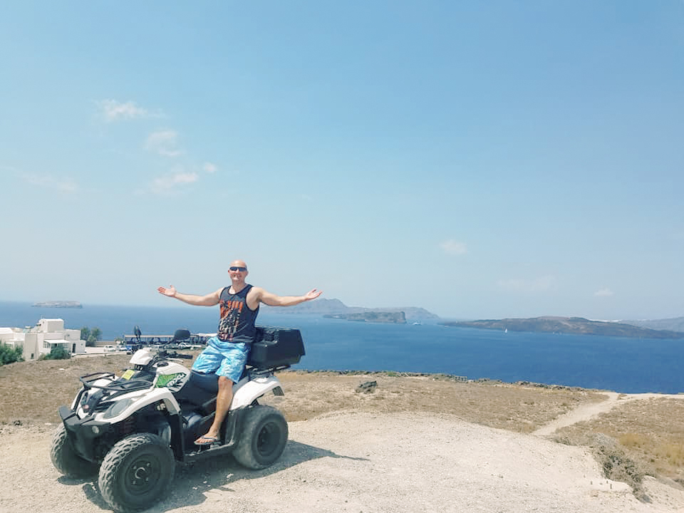 A man sits on a quad against a backdrop of the cliffside and ocean in Santorini
