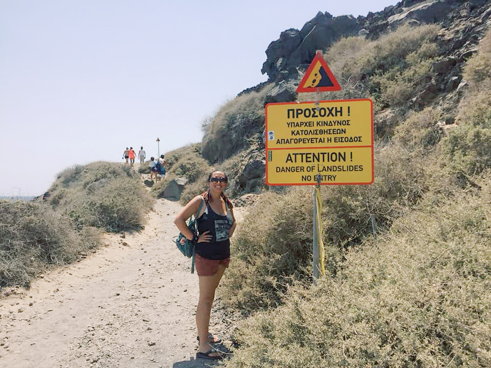 A woman poses next to a sign warning of danger on a cliffside trail in Santorini