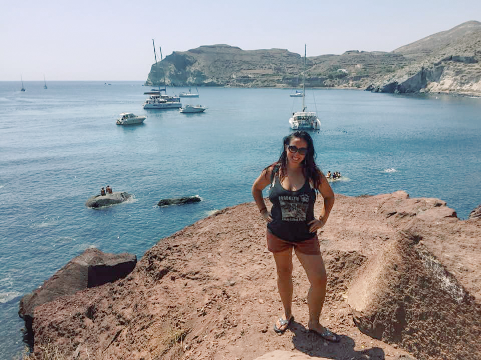 A woman poses against a backdrop of the ocean filled with boats in Santorini