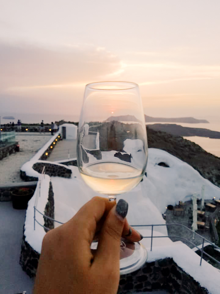 A glass of white wine held up against a backdrop of Santorini