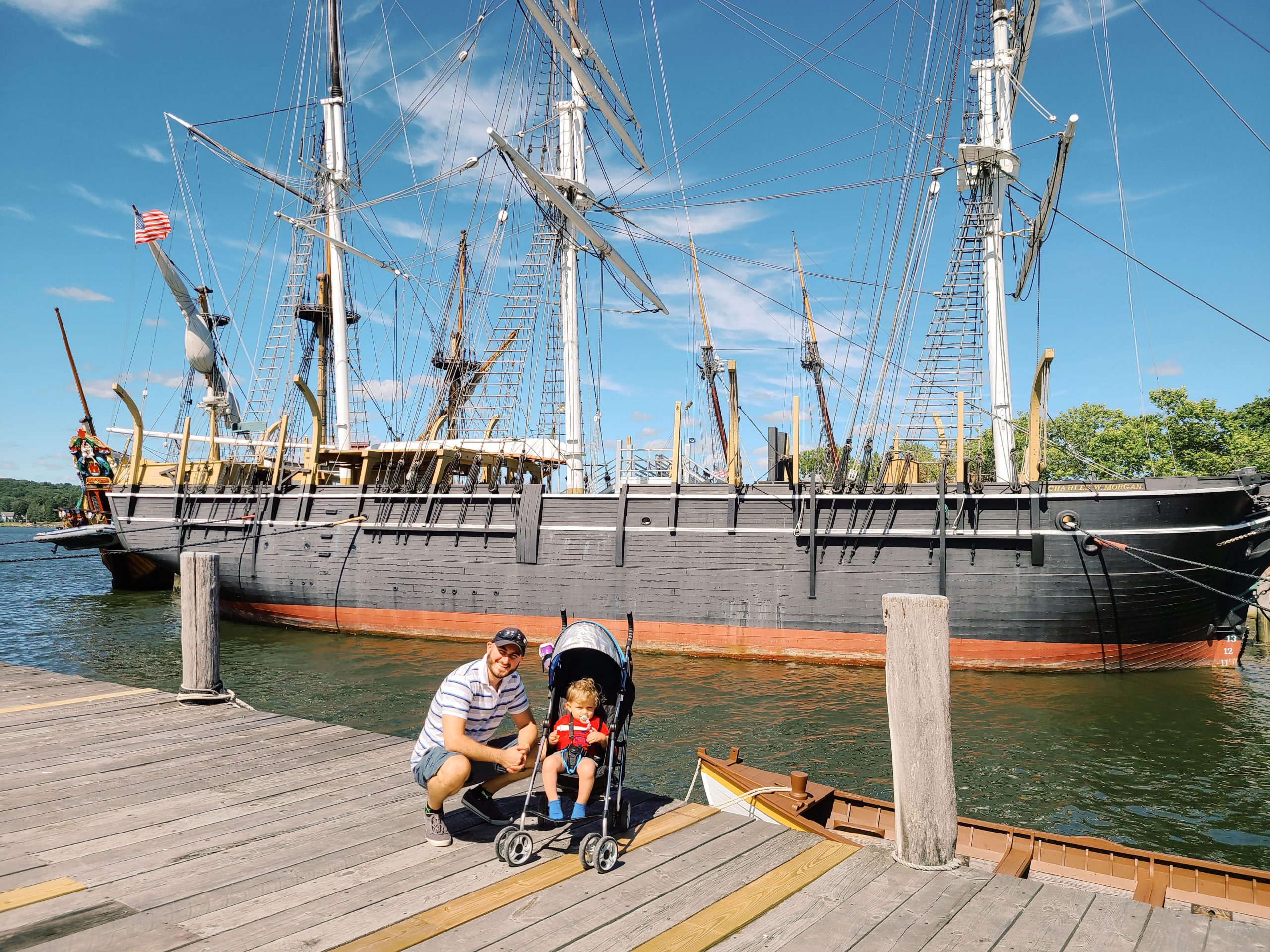 A man and child in a stroller standing on a dock posing in front of a pirate ship replica