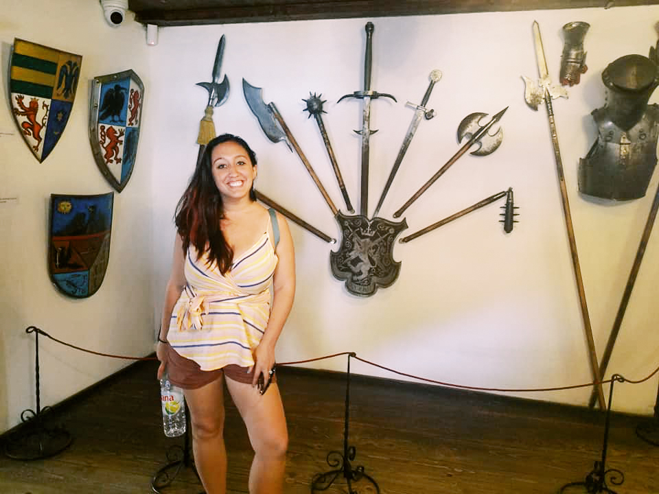 A woman poses in front of a wall of medieval shields and weapons