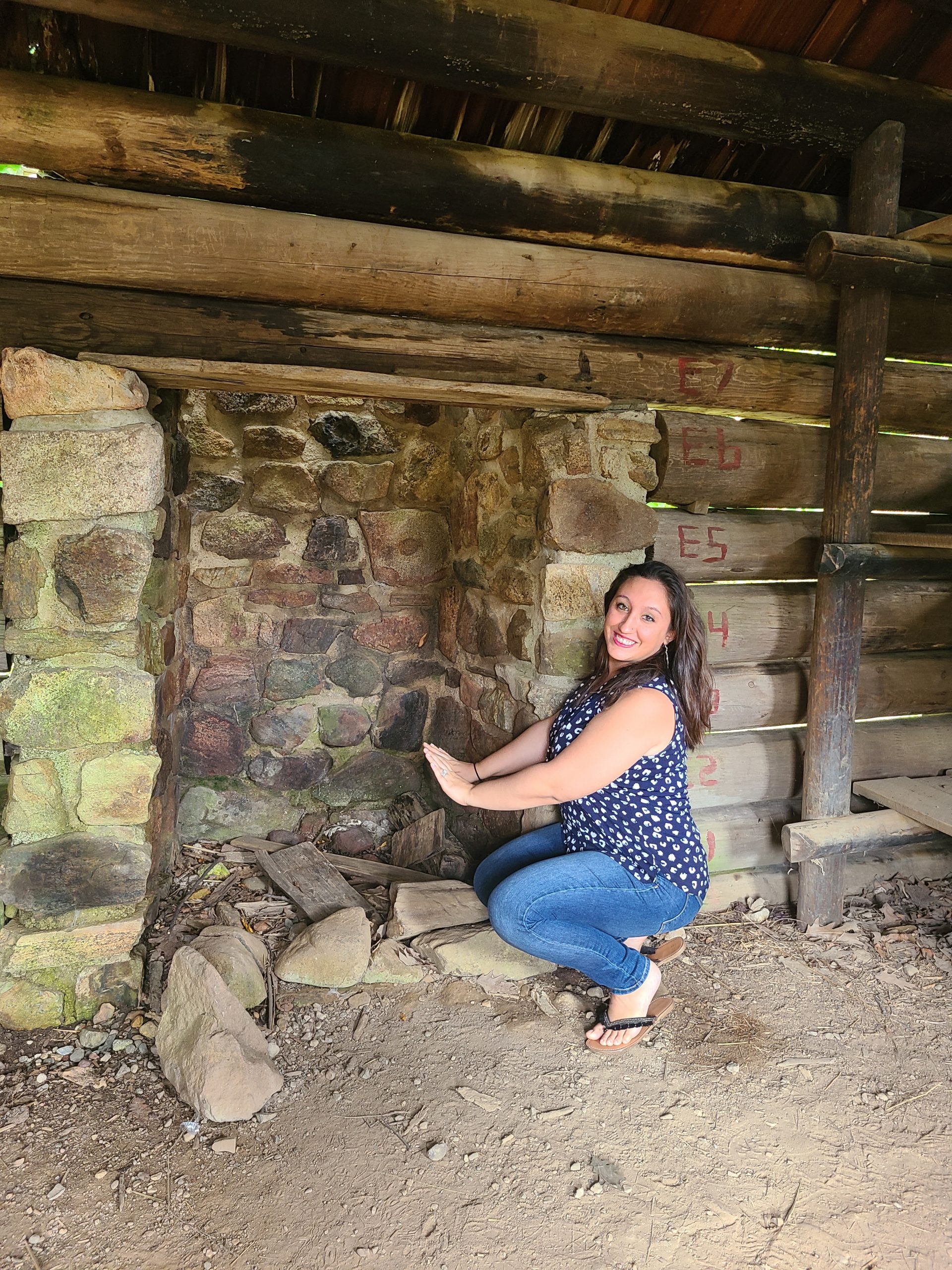 A woman sits near a hearth inside of a wooden cabin