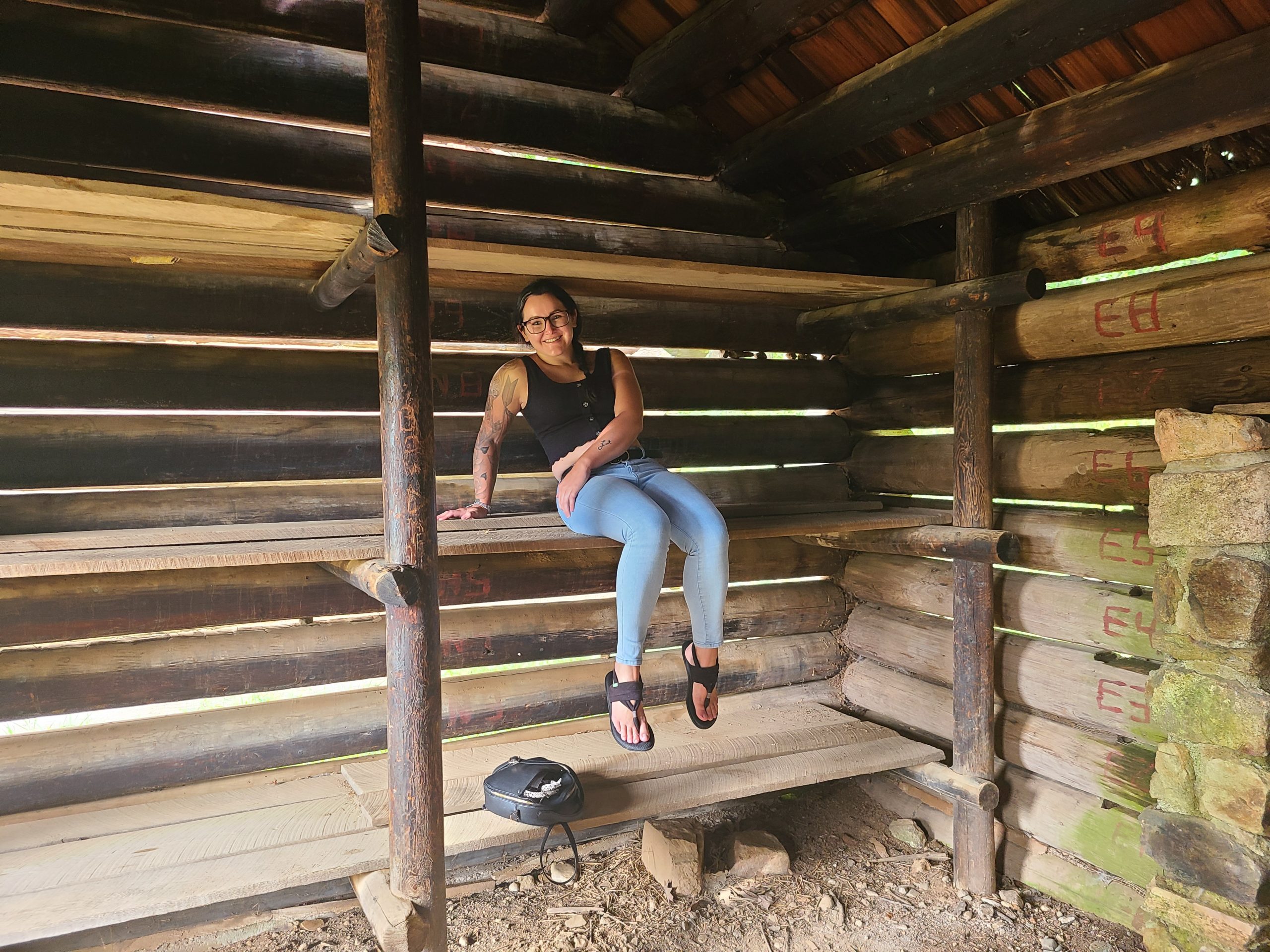 A woman sits on a wooden plank bed inside a wooden cabin