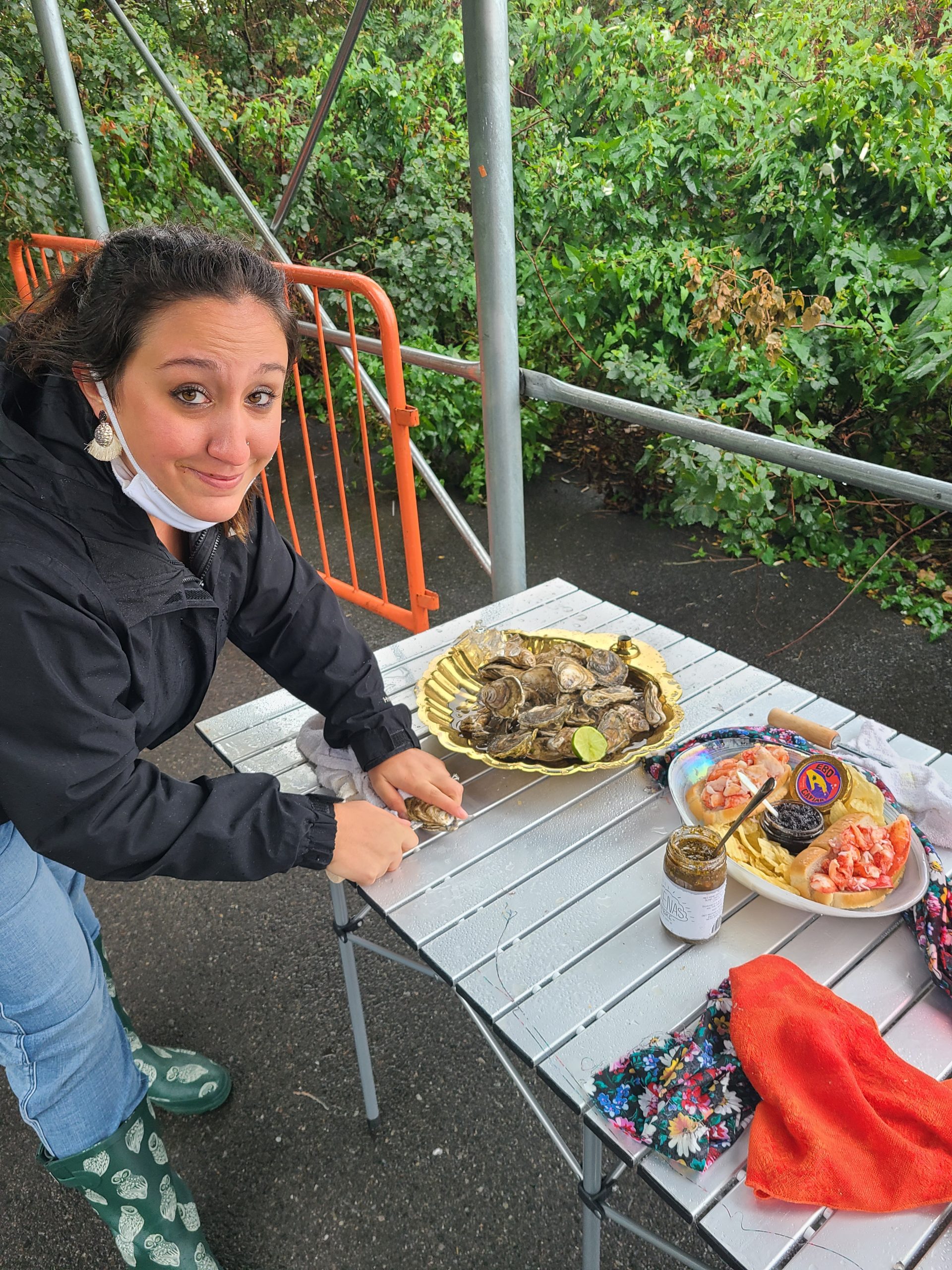 A woman shucking an oyster poses next to a table with plates of oysters and lobster rolls