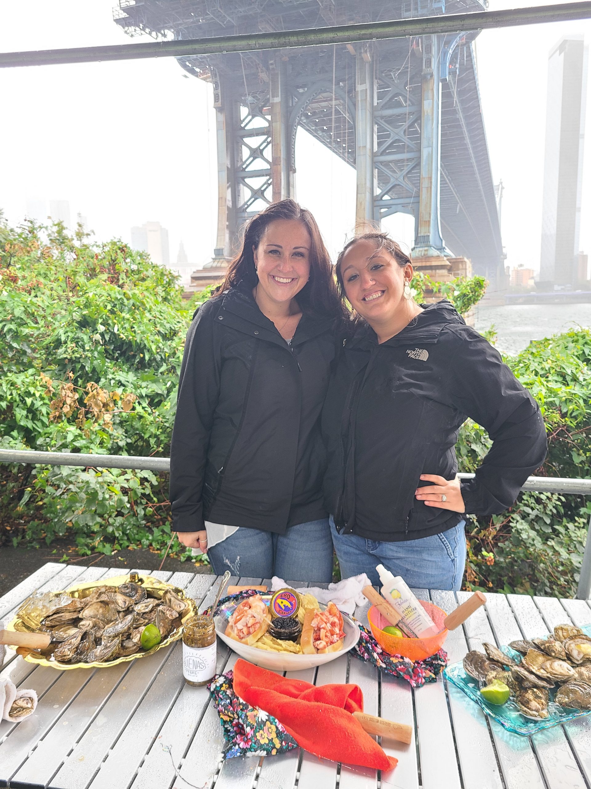 Two women pose next to a table of seafood under the Manhattan bridge
